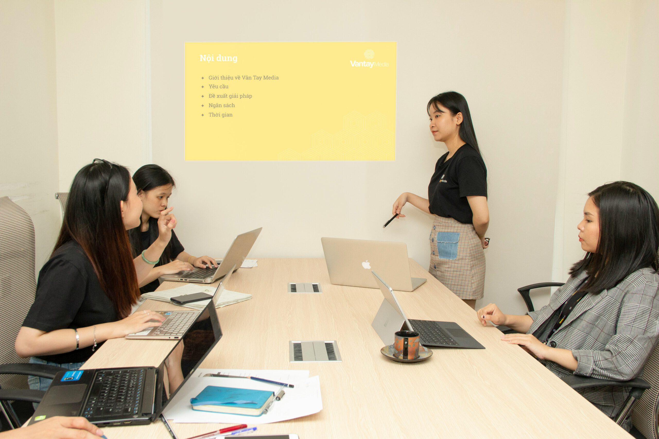 Frau steht und präsentiert vor drei sitzenden Kolleginnen mit Laptops in einem Büro – Team-Meeting und Content-Strategie-Besprechung in professionellem Arbeitsumfeld