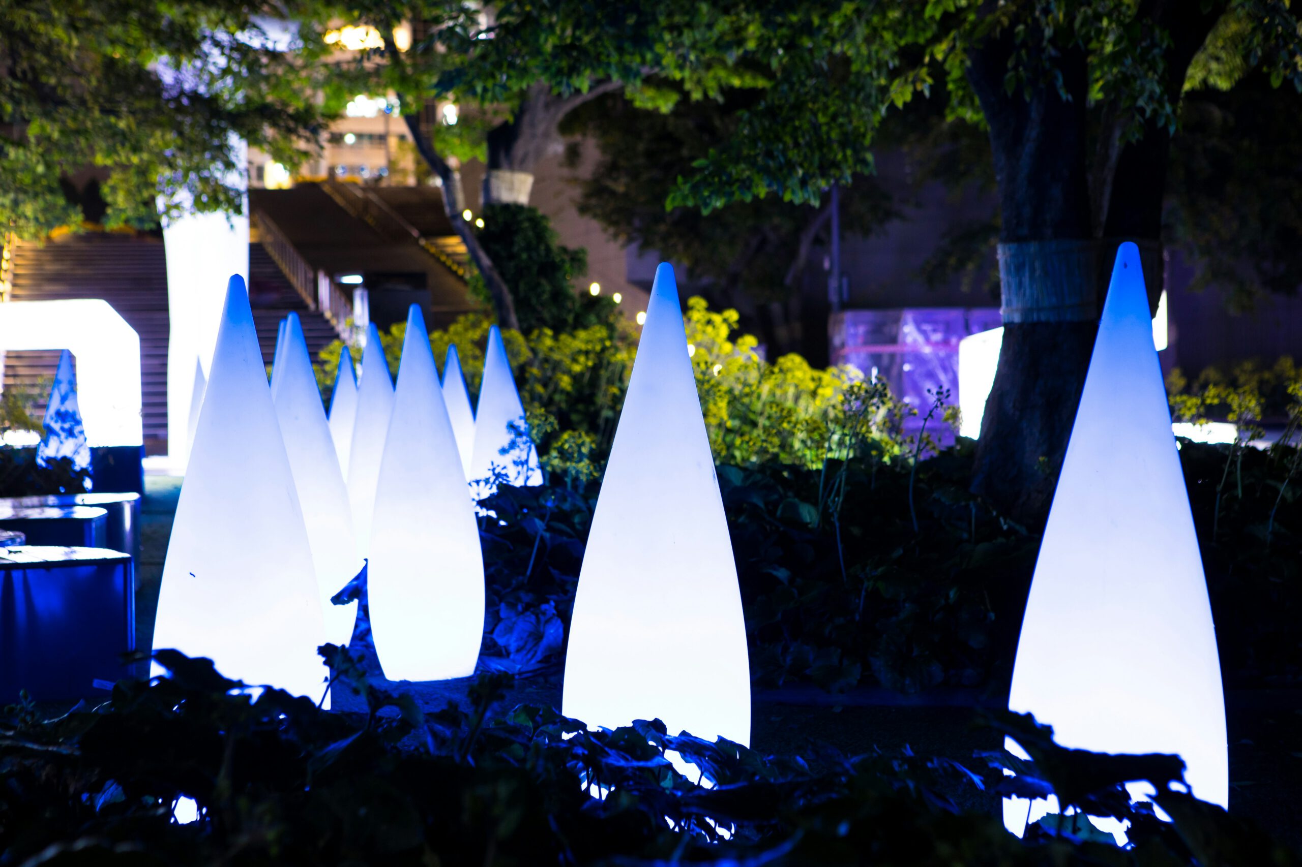 Leuchtende weiße Kegel-Lichtinstallationen in einem beleuchteten Garten bei Nacht mit Bäumen und blauem Lichtakzent – moderne Outdoor-Kunst und Lichtdesign